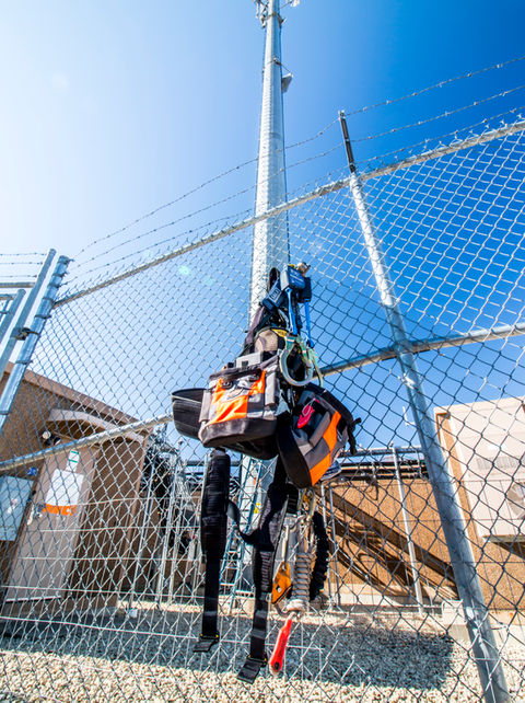 
A safety harness dangled gracefully from a fence, framed against the backdrop of a distant monopole, symbolizing the delicate balance between restraint and freedom in the realm of telecommunications infrastructure