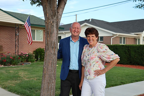 Mayor Brett Hales and his wife Cindy pose for a photo near a mature, leafy tree on a sunny day in a Murray neighborhood.