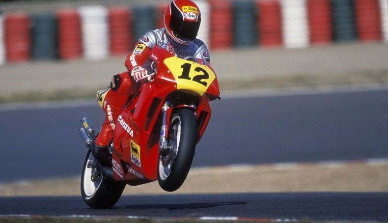 Motorcyclist in red gear pops wheelie on a red bike with number 12. Background shows blurred race track barriers in red, green, white.