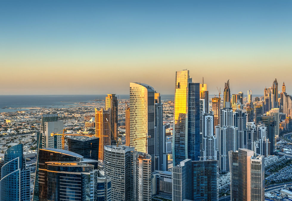 Sunlit city skyline with tall, modern skyscrapers. The ocean is visible in the background. Buildings reflect warm sunset hues.