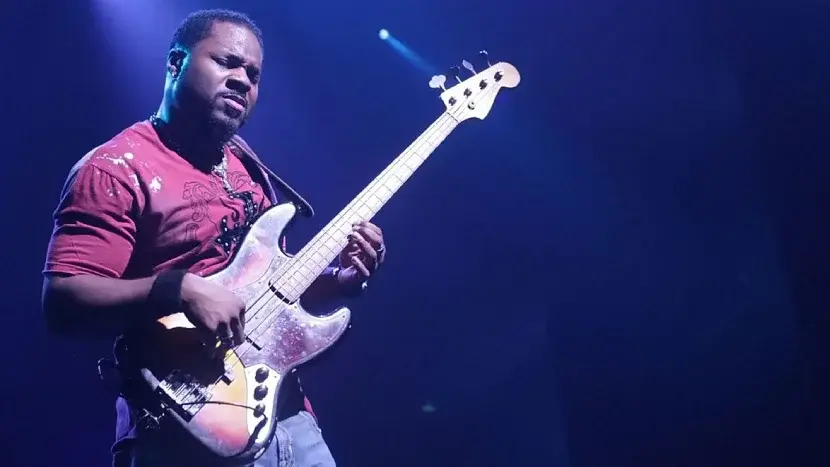 Malcolm-Jamal Warner performing on stage, playing a bass guitar under dramatic purple lighting