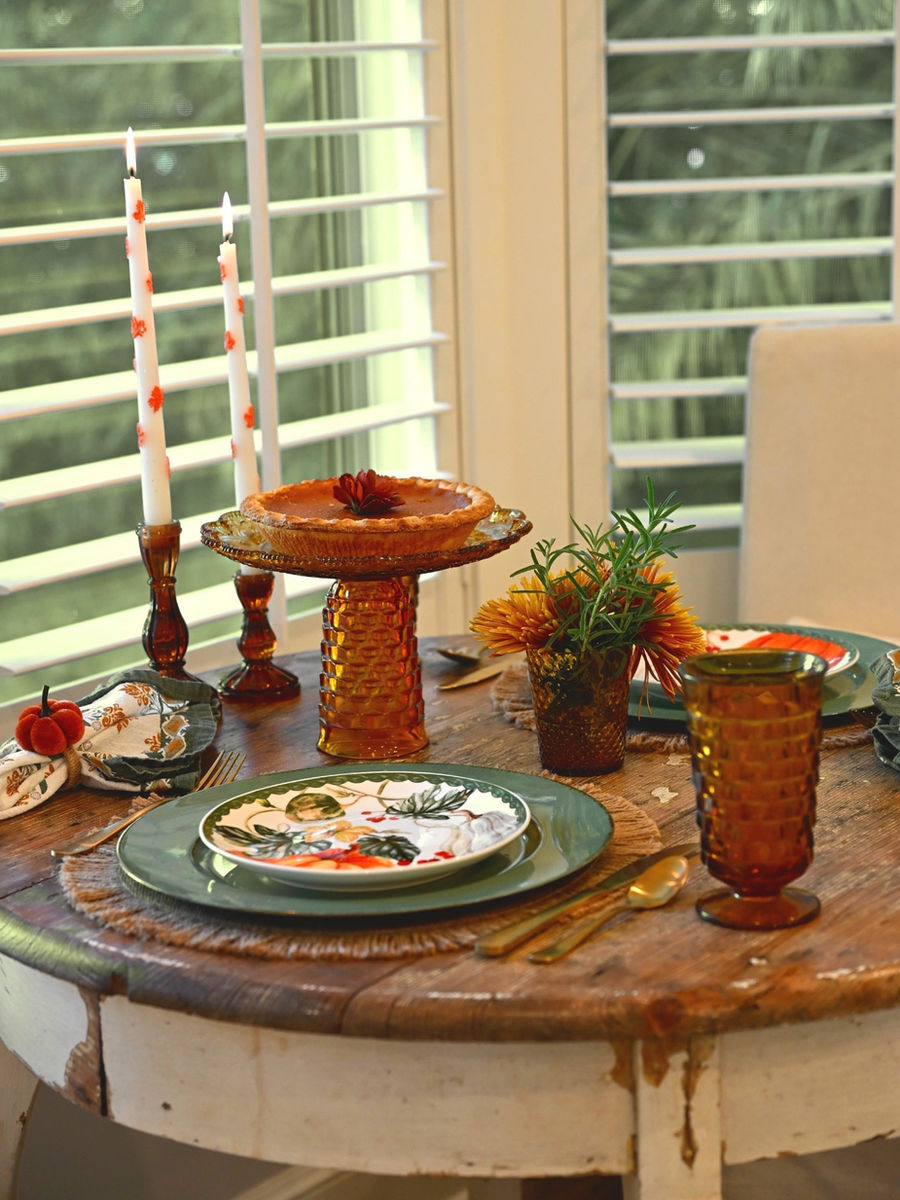 Round rustic table with full fall setting: pumpkin pie on amber cake stand, lit candles, place settings, mum vase, plantation shutters in background