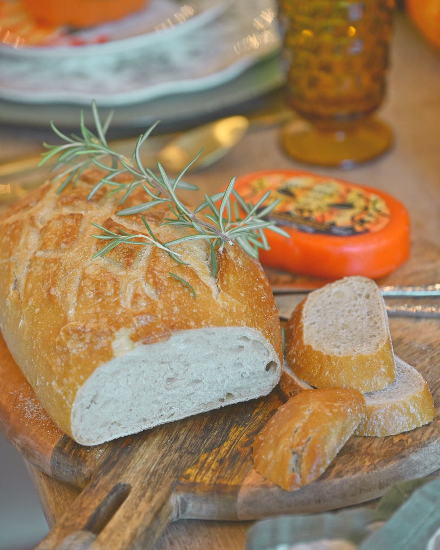 Rosemary sourdough loaf on cutting board, sliced, with pumpkin spice cheese wheel and amber glass in background