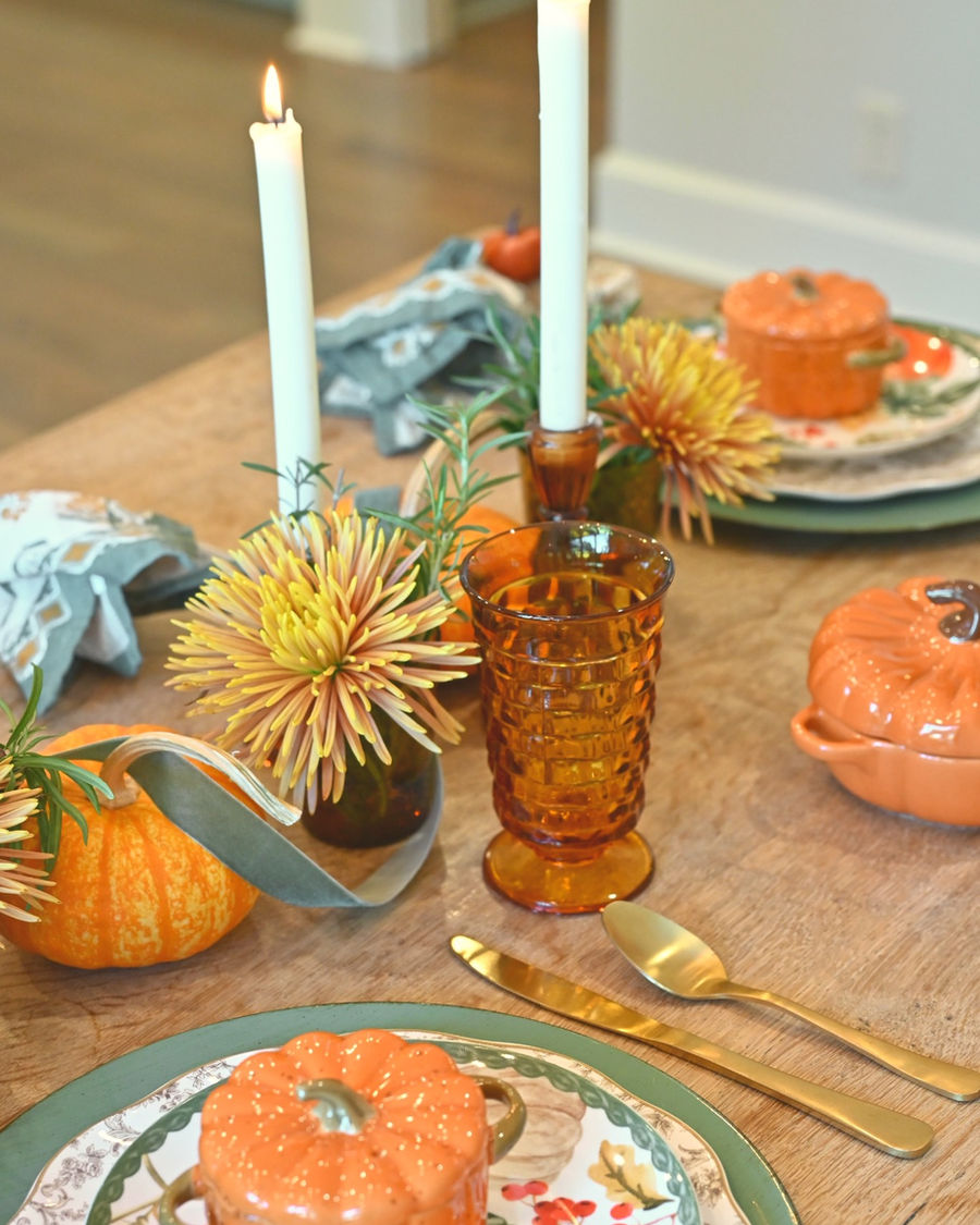Fall table side view with two lit white taper candles, amber juice glass with mum flower, place settings with pumpkin ramekins, and gold flatware