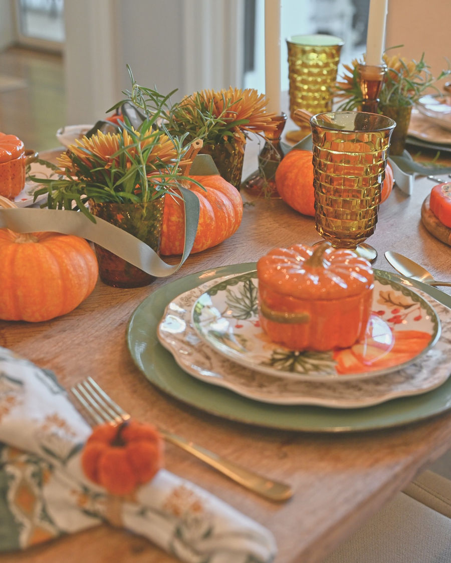 Close-up of the layered fall place setting: printed napkin with velvet pumpkin napkin ring, sage green charger, patterned plates, pumpkin ramekin on top, small pumpkins, and amber juice glass vases with mums