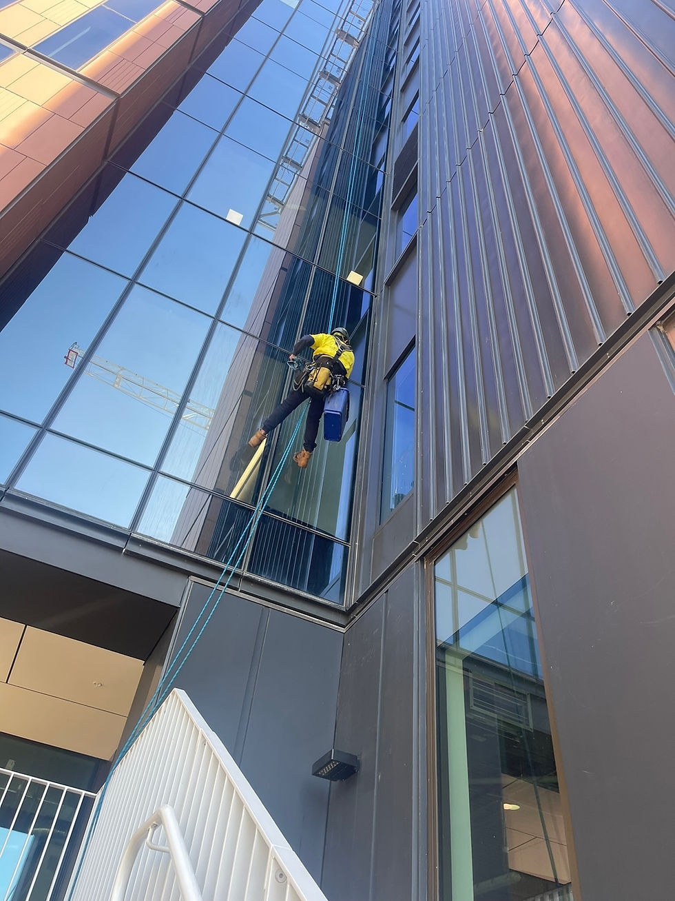 commercial window cleaning sydney: A person in yellow gear cleans windows on a tall glass building using ropes. Blue sky and crane reflection visible. Bright daylight setting.