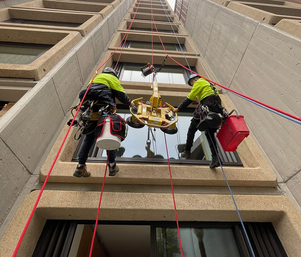 Rope access window cleaning- Two workers in safety gear clean windows on a tall building using ropes. They wear bright jackets and have equipment bags. Urban setting.