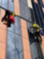 facade cleaning sydney: Two window cleaners on ropes wash a tall glass office building, one with a red bucket, against peach and dark mirrored panels.