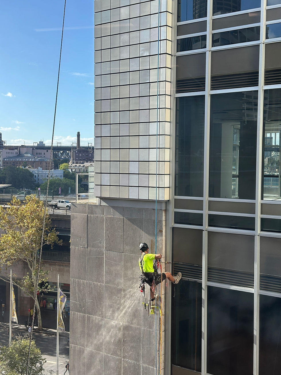 high rise window cleaning sydney- Worker in bright shirt rappels down building, cleaning windows. Urban cityscape with clear blue sky and distant structures in background.