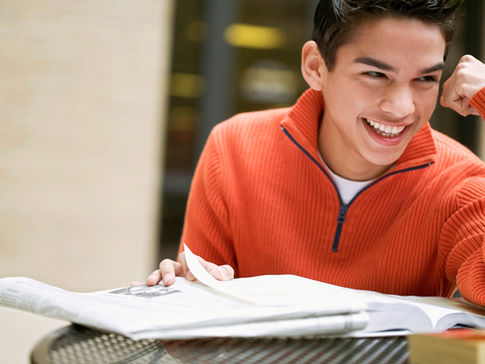 Young person in an orange sweater laughs while reading a book at a table. Bright indoor setting, joyful and relaxed mood.