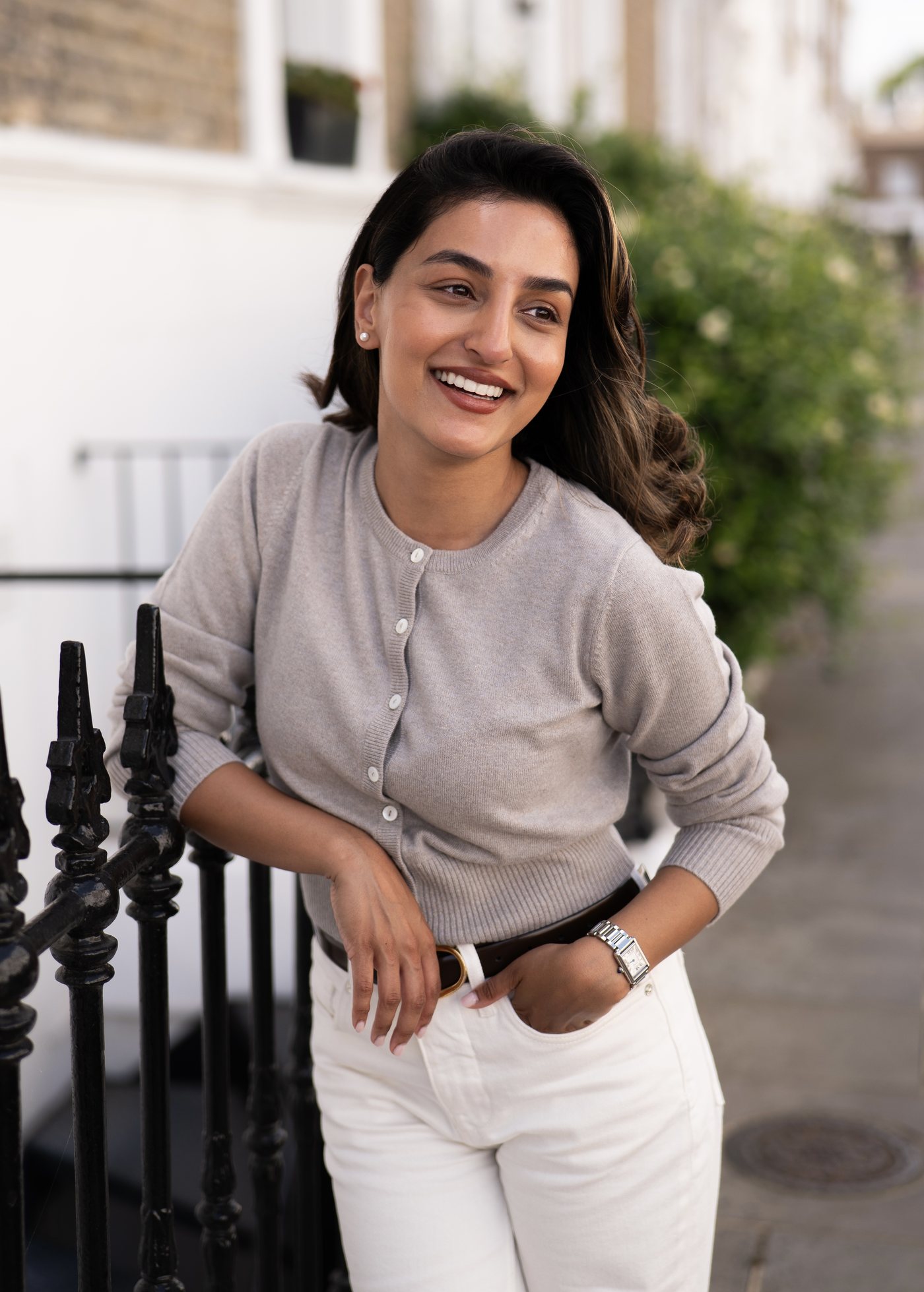 Sonam smiling by black iron railings in a grey cardigan and cream jeans