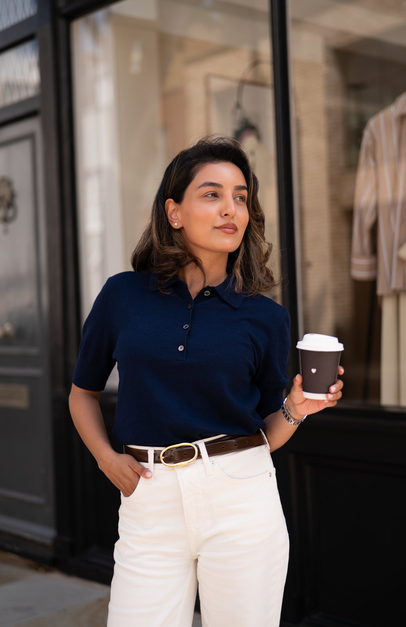 Sonam in a navy knit polo and cream jeans, holding a coffee outside a boutique