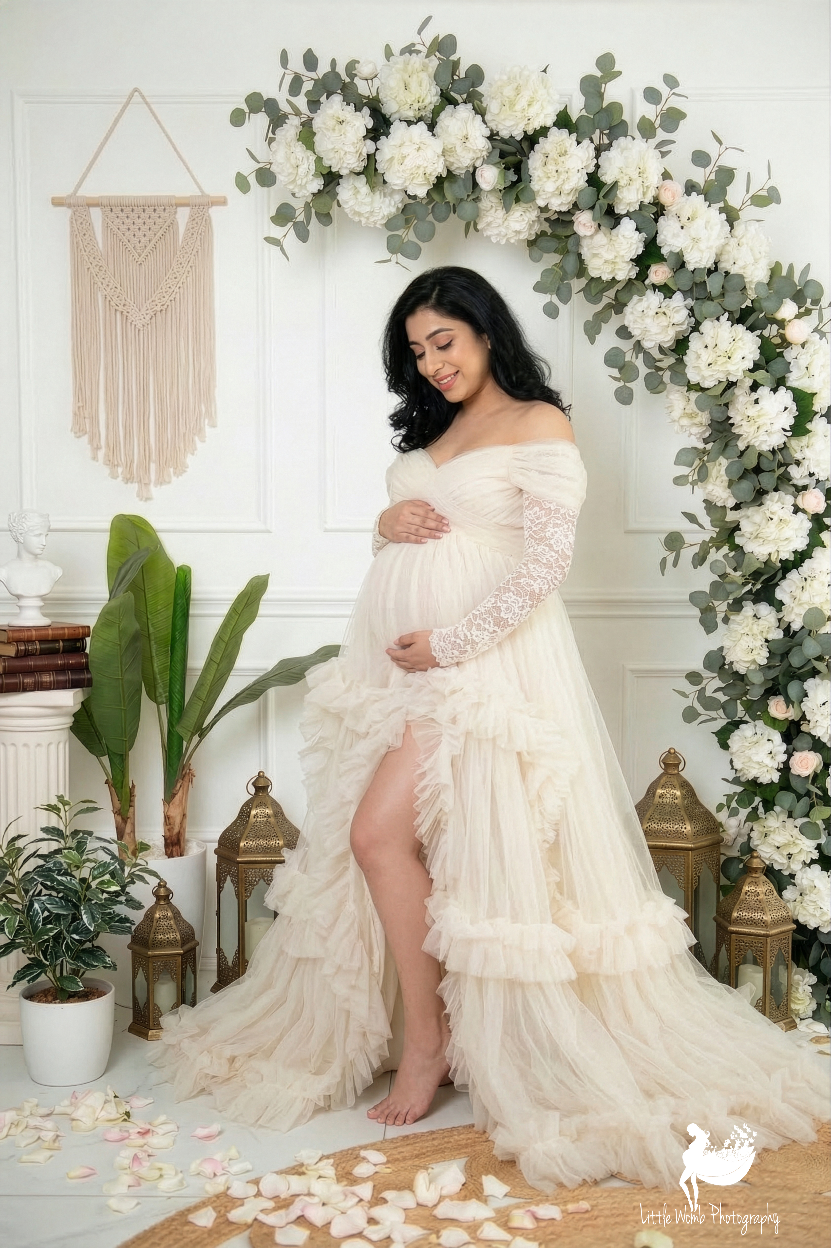 Pregnant woman posing in a white lace maternity gown under a floral archway with macrame wall art in a bright Bangalore photo studio.