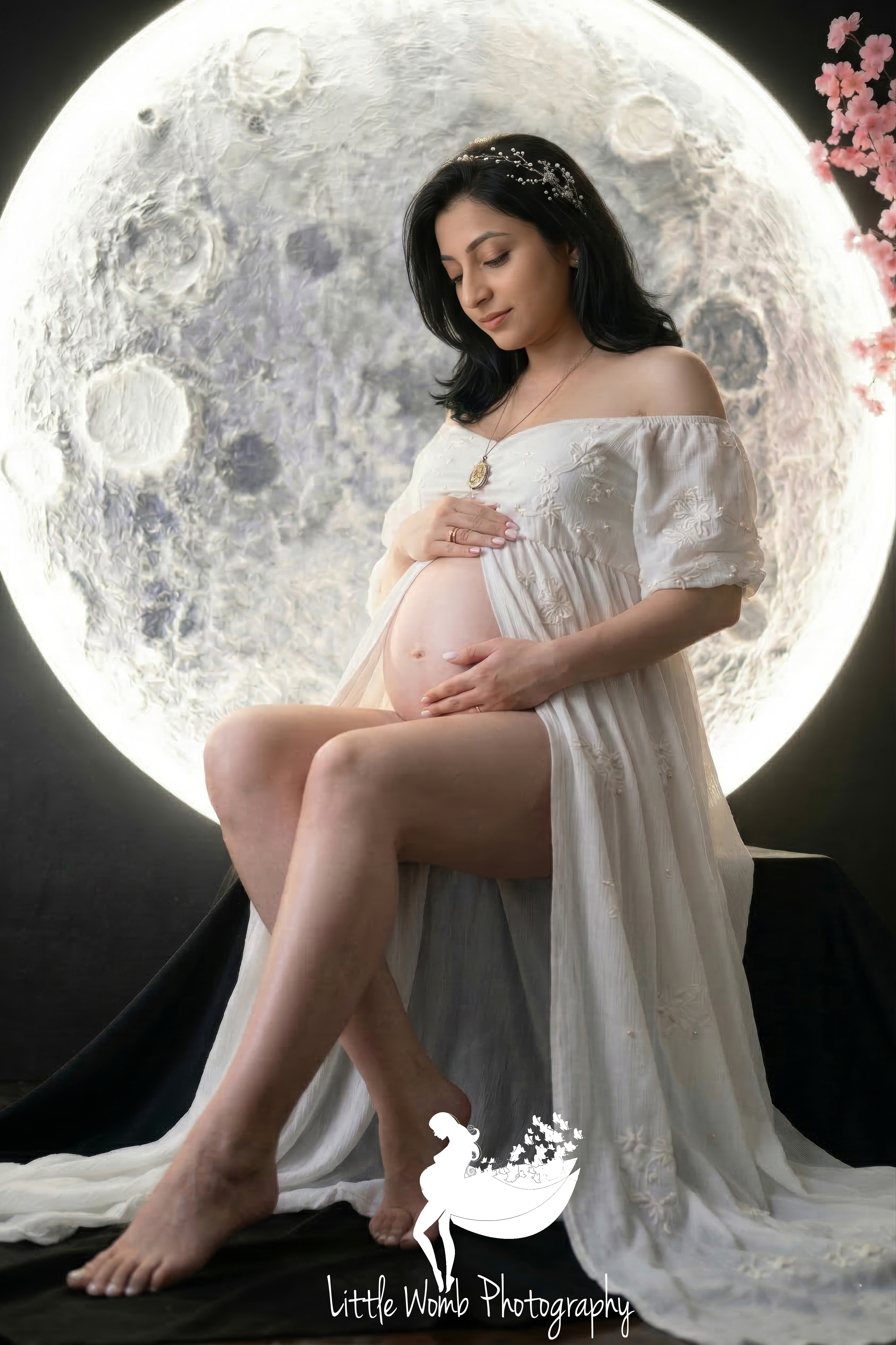 Expectant mother in a white off-shoulder gown sitting next to a large glowing full moon prop in a dark-themed creative maternity photography session in Bangalore.