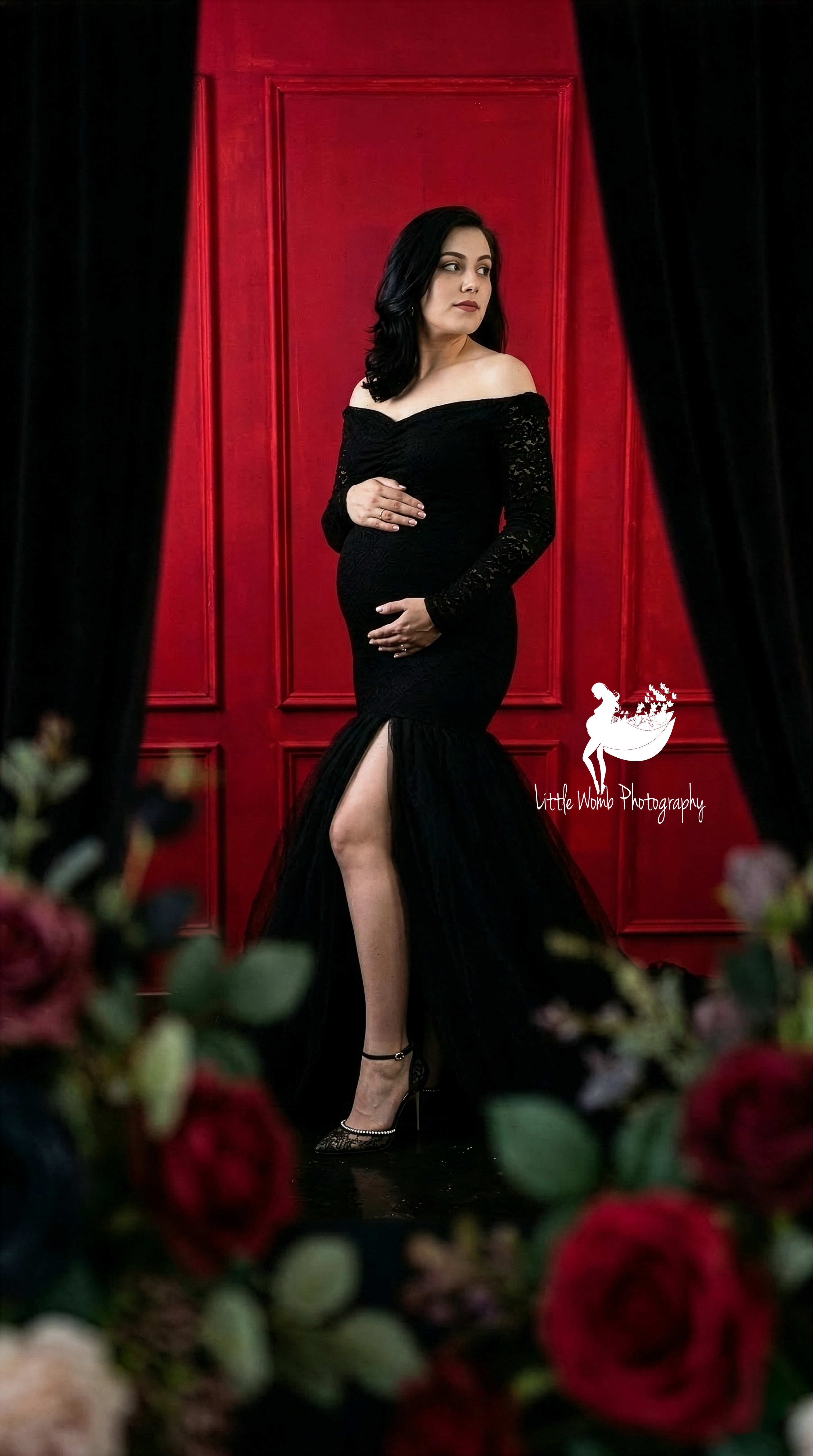 Expectant mother in a black off-shoulder gown standing against a royal red studio wall backdrop for a premium maternity photoshoot in Bangalore.