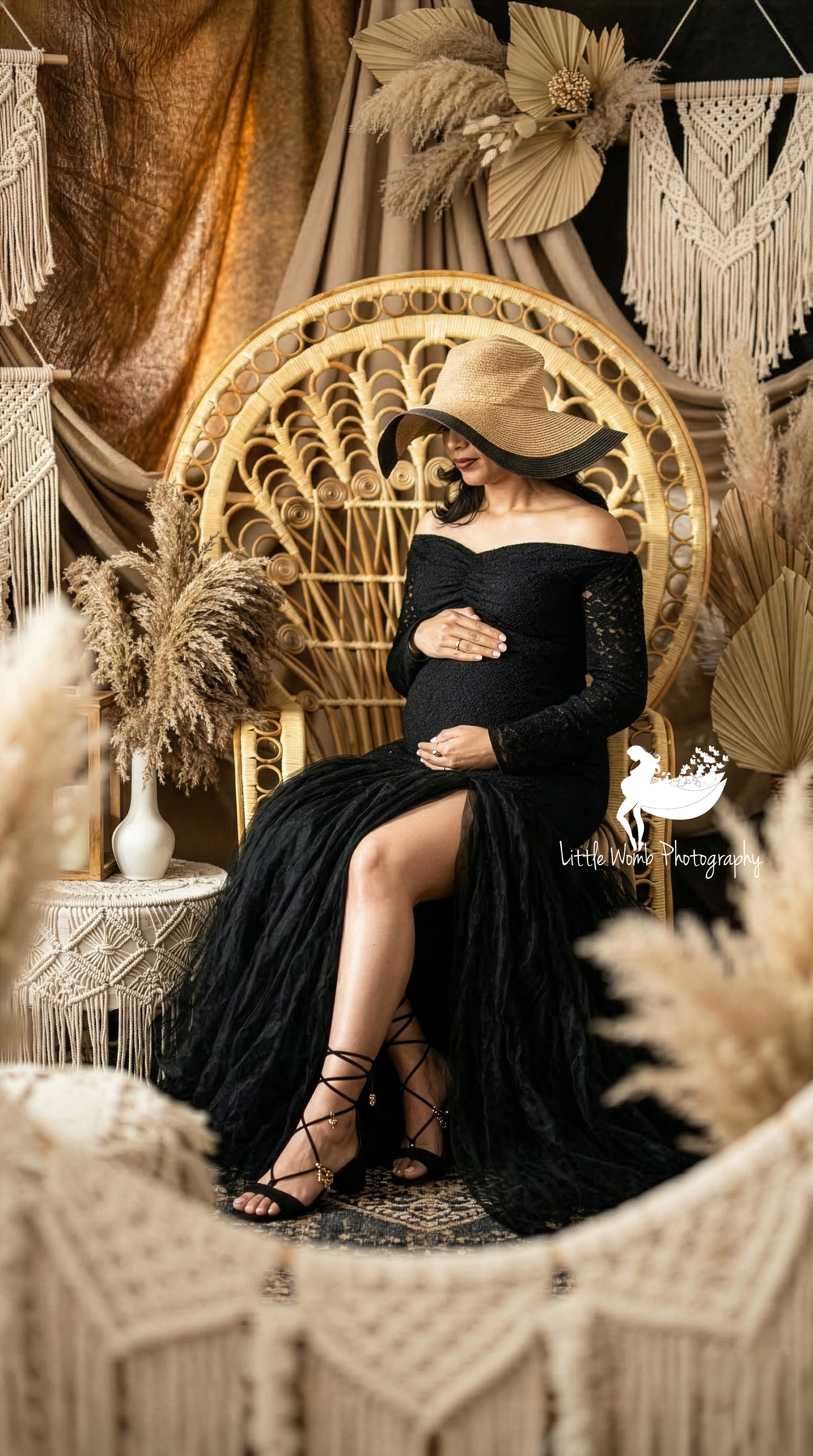 Pregnant woman in a black lace maternity gown and straw hat posing on a wicker peacock chair in a professionally decorated Bangalore photography studio.
