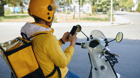 Entregador de aplicativo usando jaqueta e capacete amarelos, sentado em uma motocicleta branca, consulta o celular à beira da rua em um bairro arborizado durante o dia.