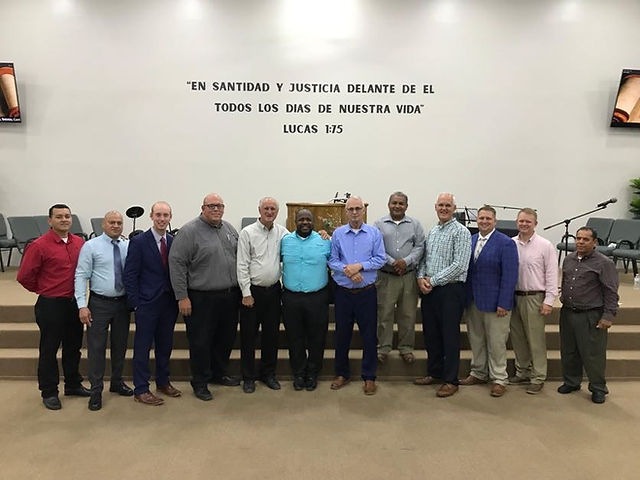 “A group of thirteen preachers standing together at the front of a church sanctuary, dressed in formal and semi-formal attire, with a Bible verse in Spanish displayed on the wall behind them.”