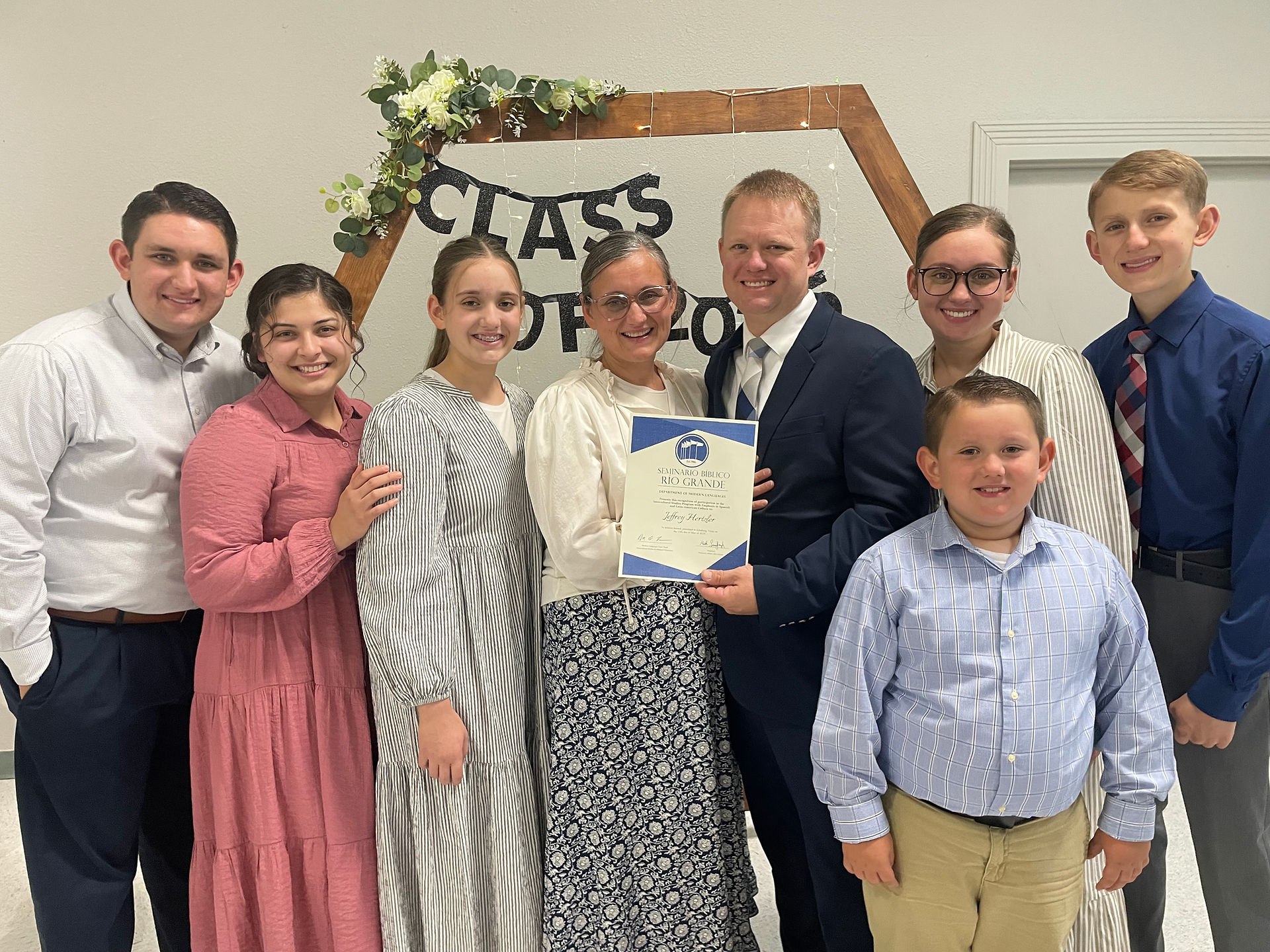 A family of eight poses together for a graduation celebration photo, with a graduate in the center holding a diploma certificate, standing in front of a 'Class of 2025' banner decorated with greenery and flowers.