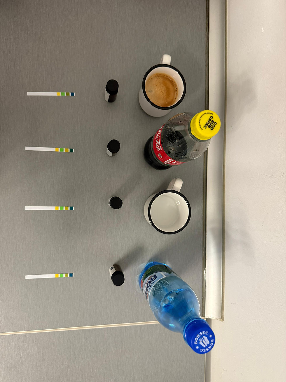 Pe masă gri: două cești albe, una cu cafea, sticle de Coca-Cola și Borsec, plus benzi de test; atmosferă de laborator.