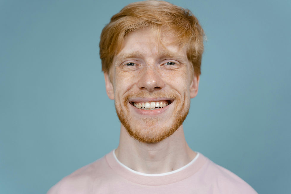 Bărbat cu păr roșcat și barbă, zâmbind fericit. Poartă un tricou roz. Fundal albastru simplu. Atmosfera este prietenoasă.