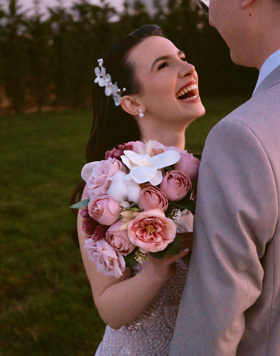 Mireasă zâmbitoare, fericită, ținând buchet de flori roz și ornament seral în păr, se uită la mirele în costum gri deschis, fundal verde.