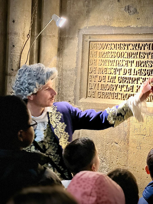 Le guide en tenue du XVIIIème siècle raconte aux enfants la légende de Pierre Perrat, l'architecte de la Cathédrale de Metz, devant son inscription funéraire.