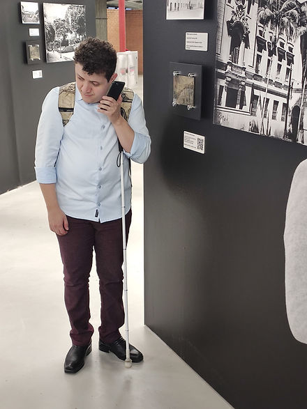 Fotografia vertical em ambiente interno de exposição. À esquerda, um homem de cabelos curtos e cacheados está em pé ao lado de um painel escuro. Traja camisa azul-clara de mangas dobradas, calça vinho escuro, sapatos pretos e usa mochila. Segura um telefone celular próximo ao ouvido esquerdo e está com uma bengala branca apoiada no no corpo. Ele está com rosto está inclinado para baixo e para a esquerda. Ao fundo e na parede ao lado dele, há fotografias em preto e branco, pequenas placas informativas com legendas e um código QR.