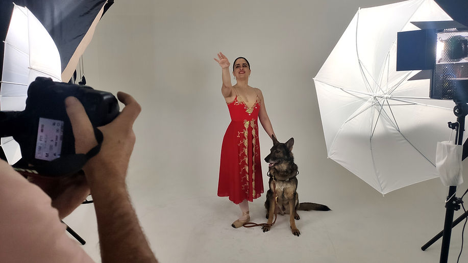 Fotografia horizontal de um estúdio fotográfico. Ao centro, uma mulher branca de cabelos escuros presos em coque, traja um vestido vermelho e sapatilhas clara. Ela posa ao lado de um cão sentado, com um dos braços levantado. À frente dela e à esquerda, um fot