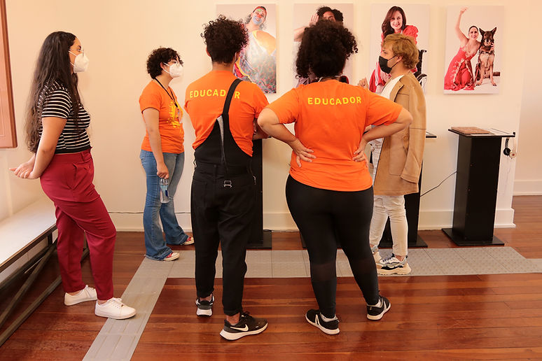 Ao centro, cinco pessoas conversam diante de fotografias expostas na parede. Três delas trajam camisetas laranja com a palavra “EDUCADOR” nas costas. Há piso tátil no chão e totens expositivos com peças táteis e fones de ouvido junto à parede.