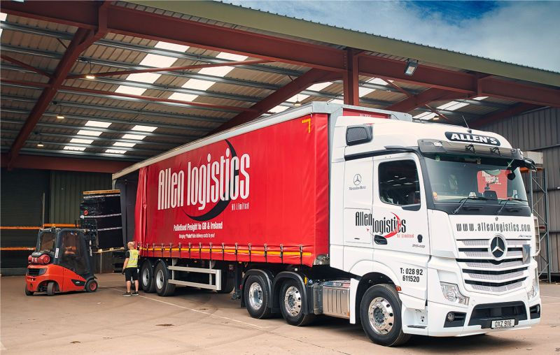 Red truck labeled "Allen Logistics" parked in a warehouse. A person operates a forklift moving goods. Bright and busy setting.