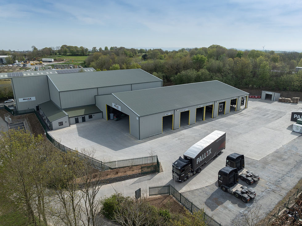 Aerial view of a logistics facility with trucks labeled "PALLEX" near loading bays. Gray buildings and a forested background under a blue sky.