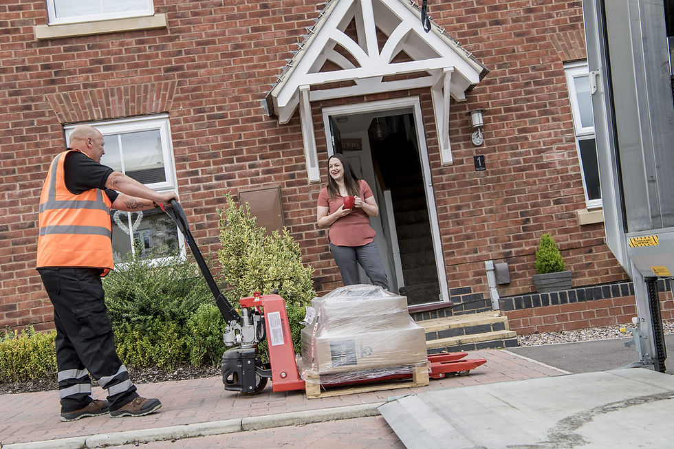 A man unloading a pallet from the back of a truck outside a house while a woman waits outside the door.