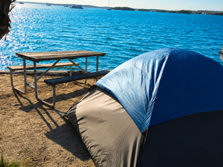 Blue and black tent beside a picnic table on a sandy beach, with bright blue water, boats, and distant islands under clear sky