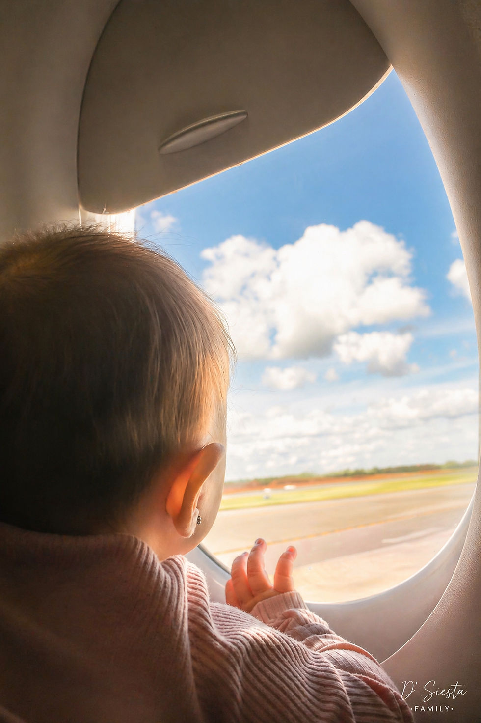 A baby in a pink sweater gazes out an airplane window at a sunny sky and clouds. The mood is curious and peaceful.