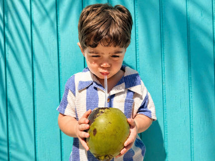 Young boy in blue checkered outfit drinks from a coconut with a straw against a bright turquoise wall, shadows of palm leaves above.