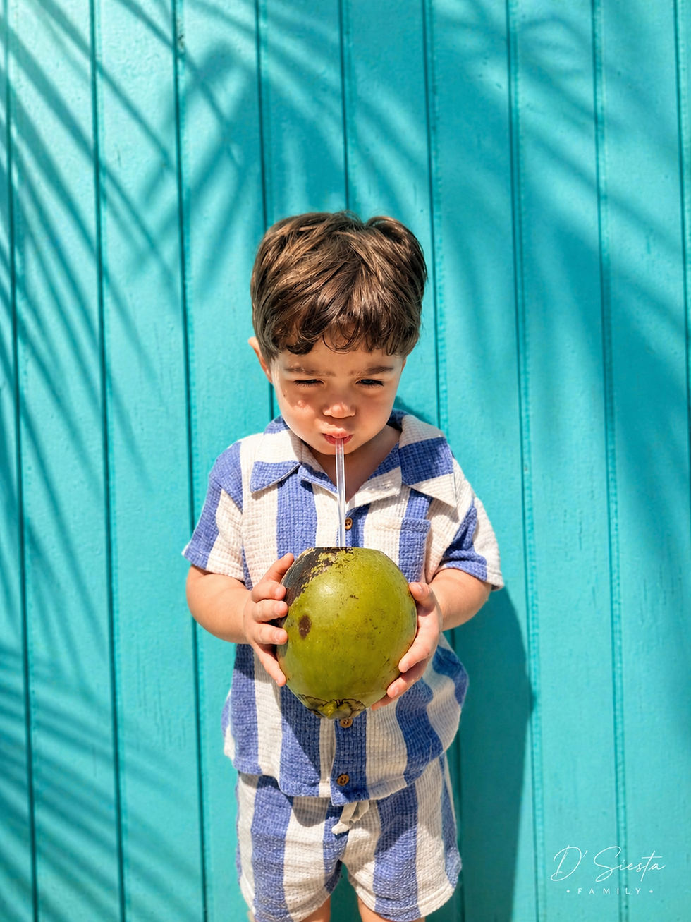 Young boy in blue checkered outfit drinks from a coconut with a straw against a bright turquoise wall, shadows of palm leaves above.