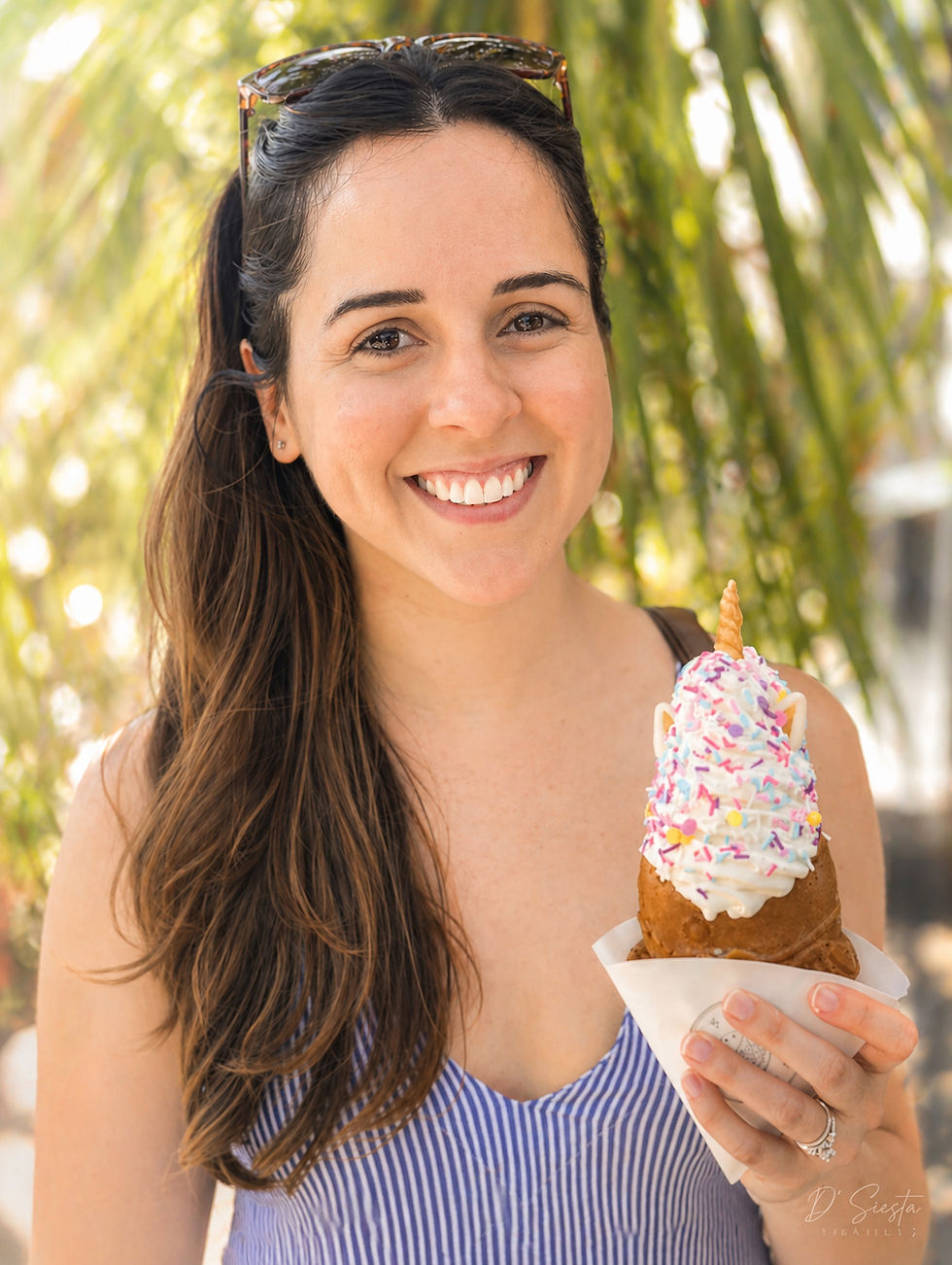 Smiling woman holding a dessert cone topped with colorful sprinkles. She's outdoors with green foliage in the background, wearing a striped top.