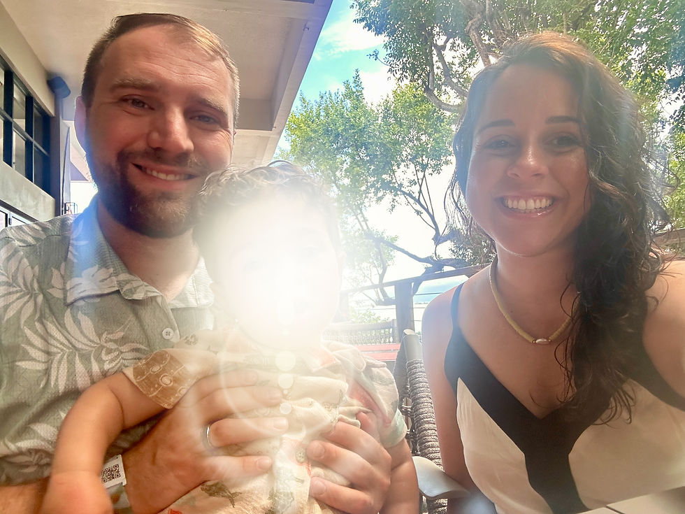 Smiling couple and child outdoors, sitting at a table. Bright daylight, with trees and blue sky in the background. Sunny and cheerful mood.