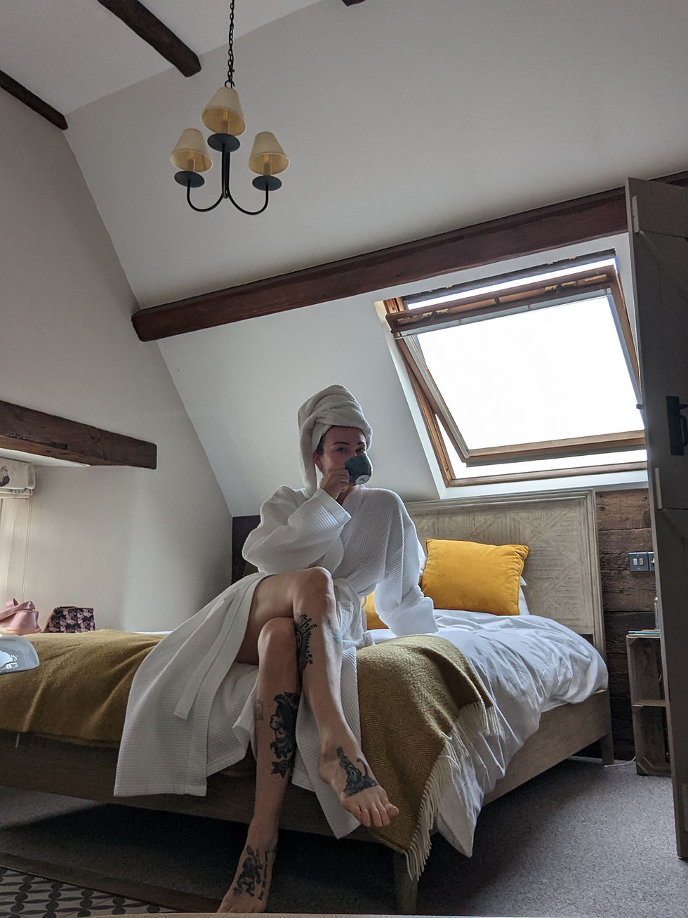 Person in a white robe and towel sits on a bed, drinking from a mug. Room has a rustic vibe with wood beams, yellow cushion, and skylight.