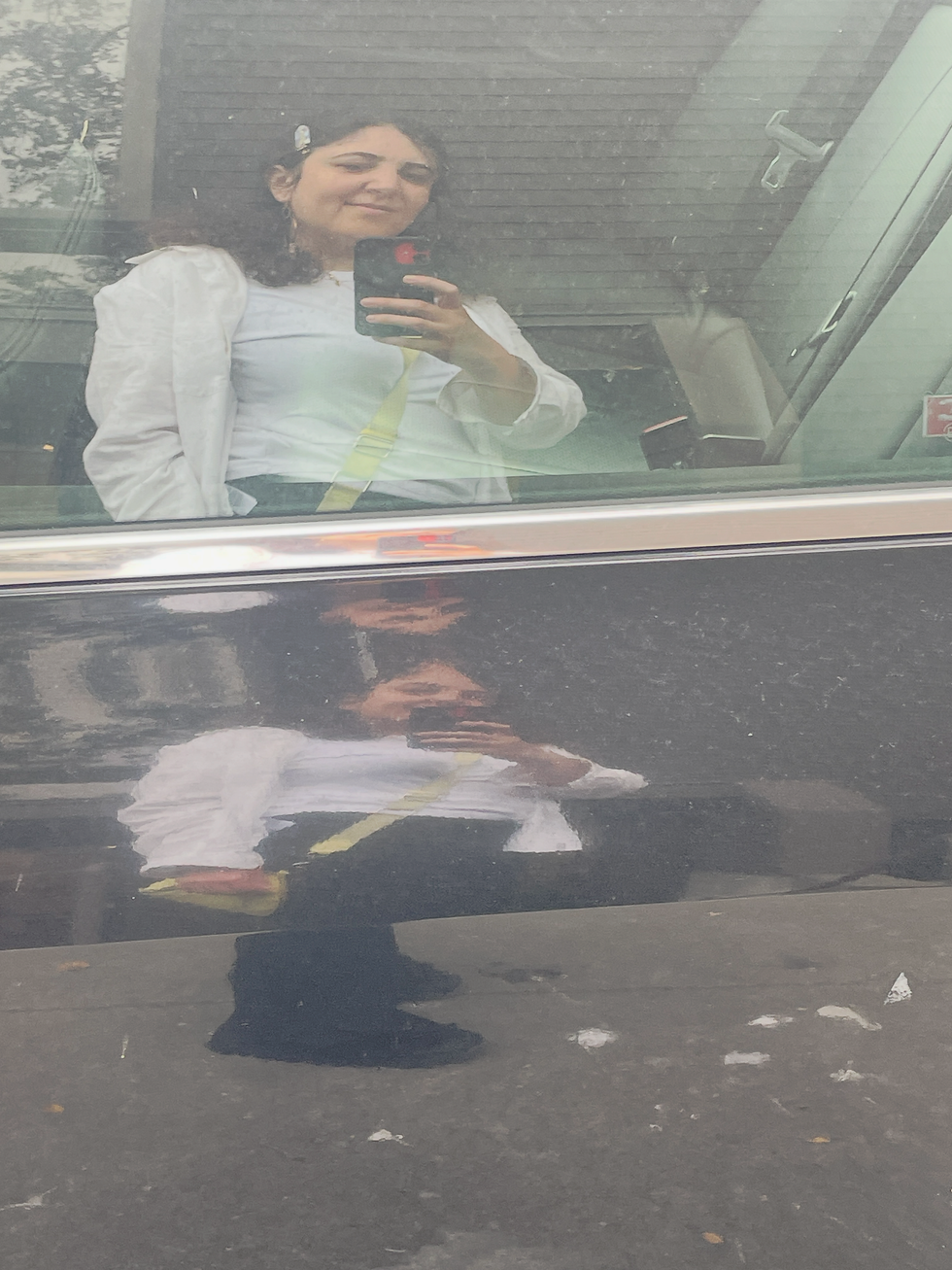 Woman in white top takes a mirror selfie in a dusty car window reflection, with a faint smile and dark interior background.