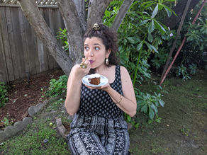 Woman in patterned black outfit eats molasses cake from a plate in a leafy garden, looking playful and surprised.
