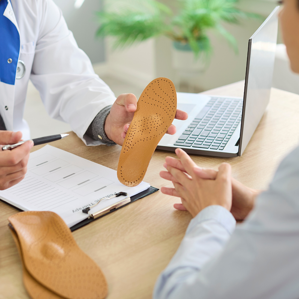 Doctor shows a tan orthotic insole to a patient at a desk with laptop and clipboard in a bright clinic.