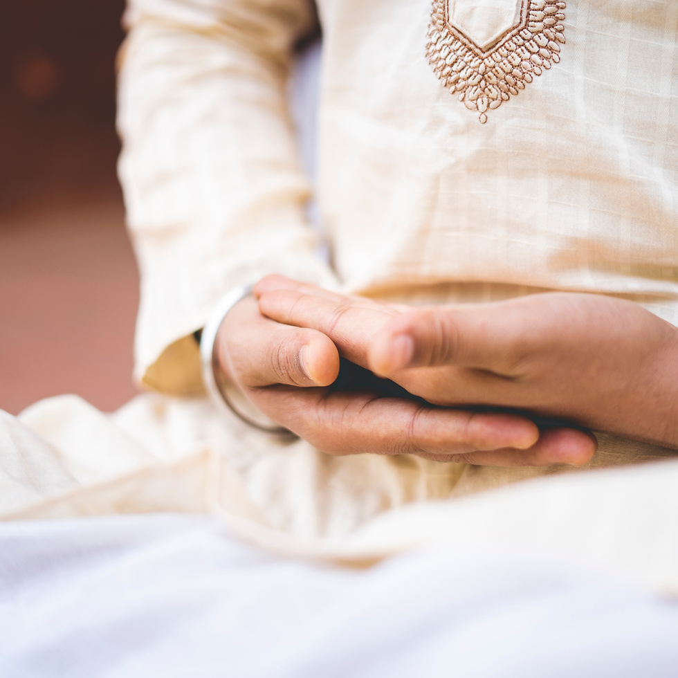 Close-up of folded hands resting in a lap, with a person in cream embroidered clothing and a calm, reflective mood.