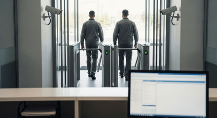 Entrada empresarial com catracas de controle de acesso, dois colaboradores passando e monitor de sistema ao fundo, representando governança baseada em registro físico estruturado.