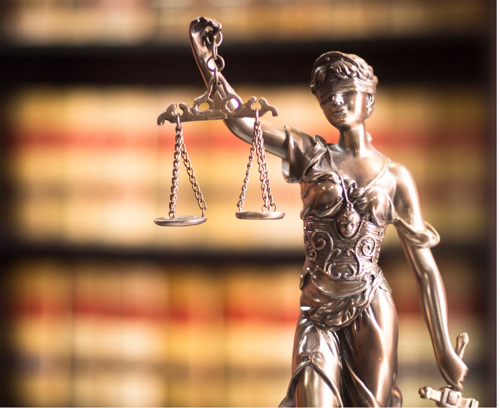 Bronze Lady Justice statue holding scales, blindfolded, stands against blurred bookshelves. Dim light emphasizes balance and fairness.