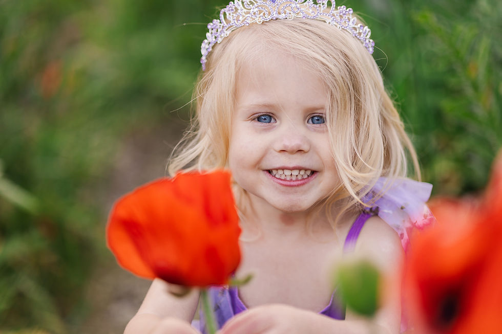 A smiling child with a tiara holds a bright red flower, wearing a purple outfit, surrounded by greenery. Joyful and vibrant scene.