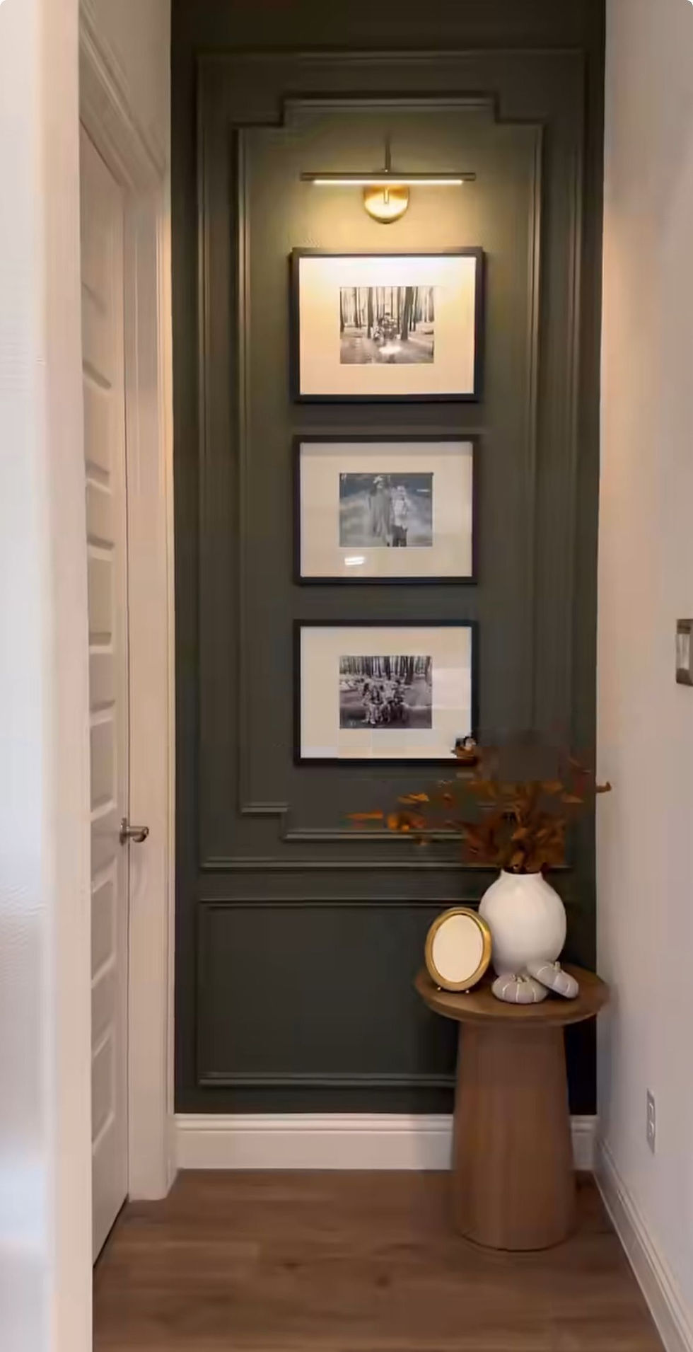 Three black-and-white photos on a dark green wall, lit from above. A small wooden table with a white vase and dried leaves. Cozy ambiance.
