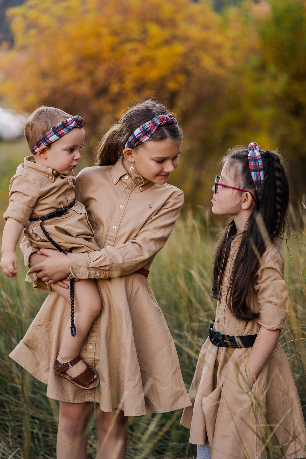 Three children in tan dresses and plaid headbands stand in grassy field, smiling at each other with autumn trees in the background.