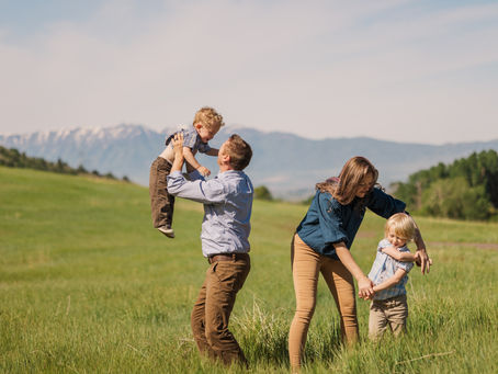 Family playing in a sunny meadow with mountains in the background; a man lifts a child, while a woman holds hands with another child.